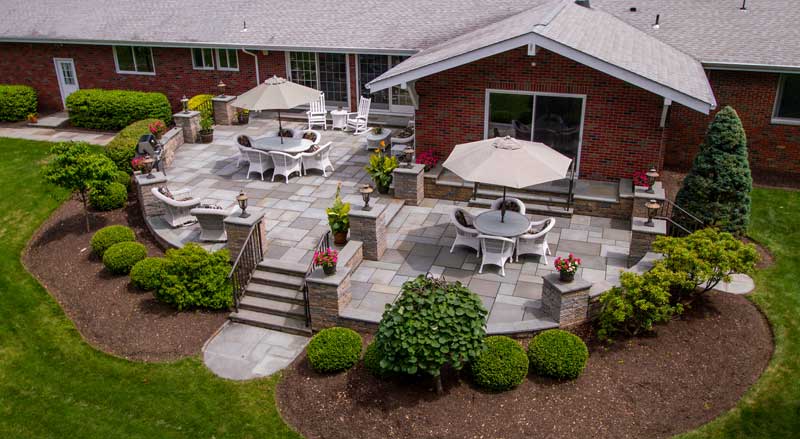 A stylish stone patio with seating areas and umbrellas, bordered by well-trimmed shrubs and precisely edged garden beds.