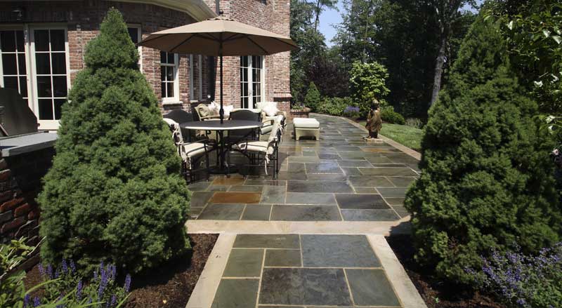 Elegant stone patio with outdoor dining set, umbrella, and manicured evergreens creating a cozy outdoor retreat.