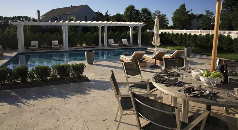 Luxury poolside hardscaping featuring a stone patio, a modern dining area, and a white pergola with lounge seating.