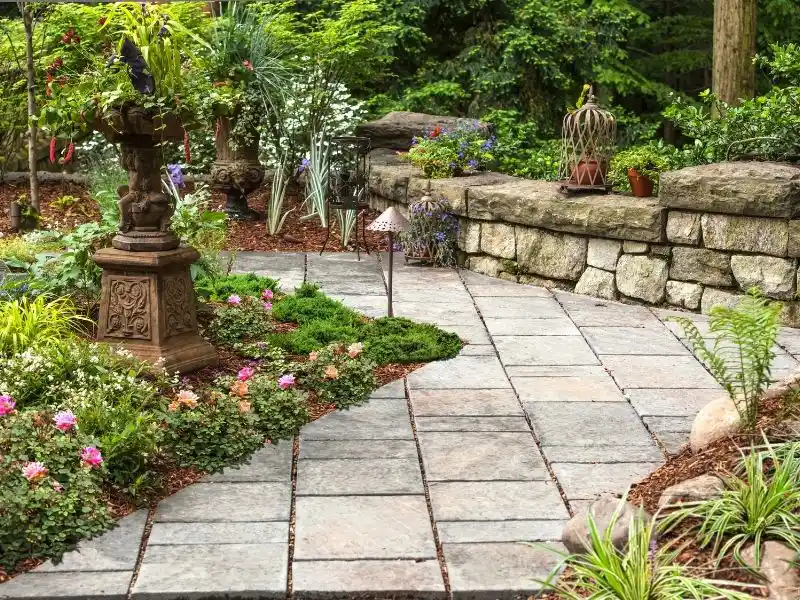 Stone garden path with birdbath, flowers, and lush greenery beside a rustic wall
