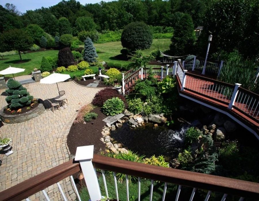Backyard garden with a paved patio, tables with umbrellas, a wooden deck, and a small pond surrounded by plants