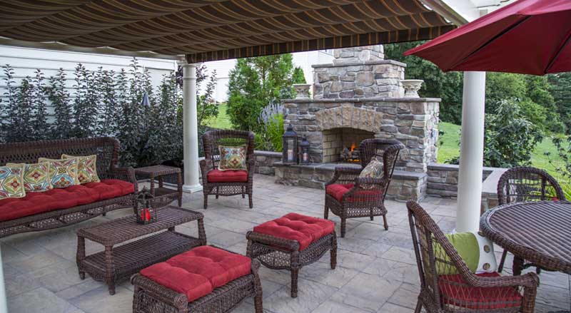 Covered outdoor living patio with stone fireplace, comfortable seating, and professionally landscaped surroundings designed for spring entertaining in Morris County, NJ.