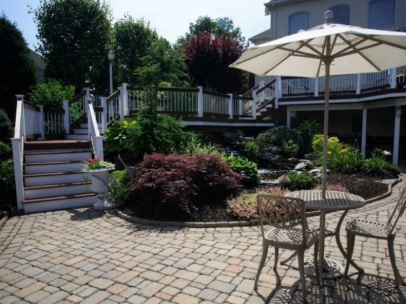 Paver patio with umbrella seating, landscaped garden, and elevated deck
