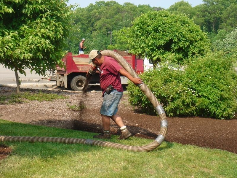 Worker spreading mulch with a hose from a truck in a landscaped garden.
