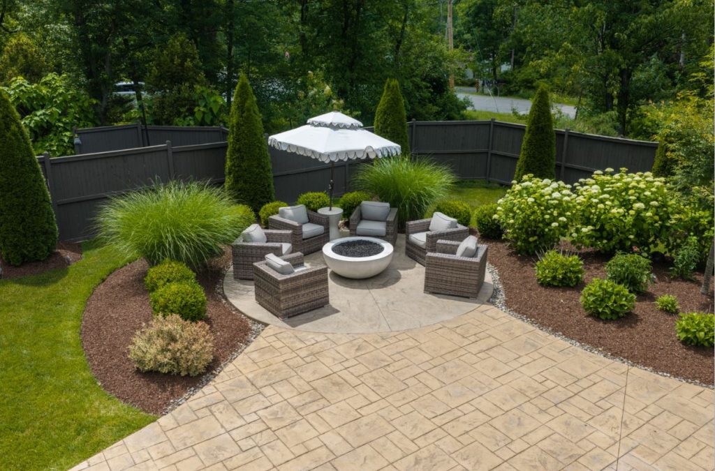Outdoor patio with wicker chairs, fire pit, white umbrella, shrubs, and landscaped garden beds