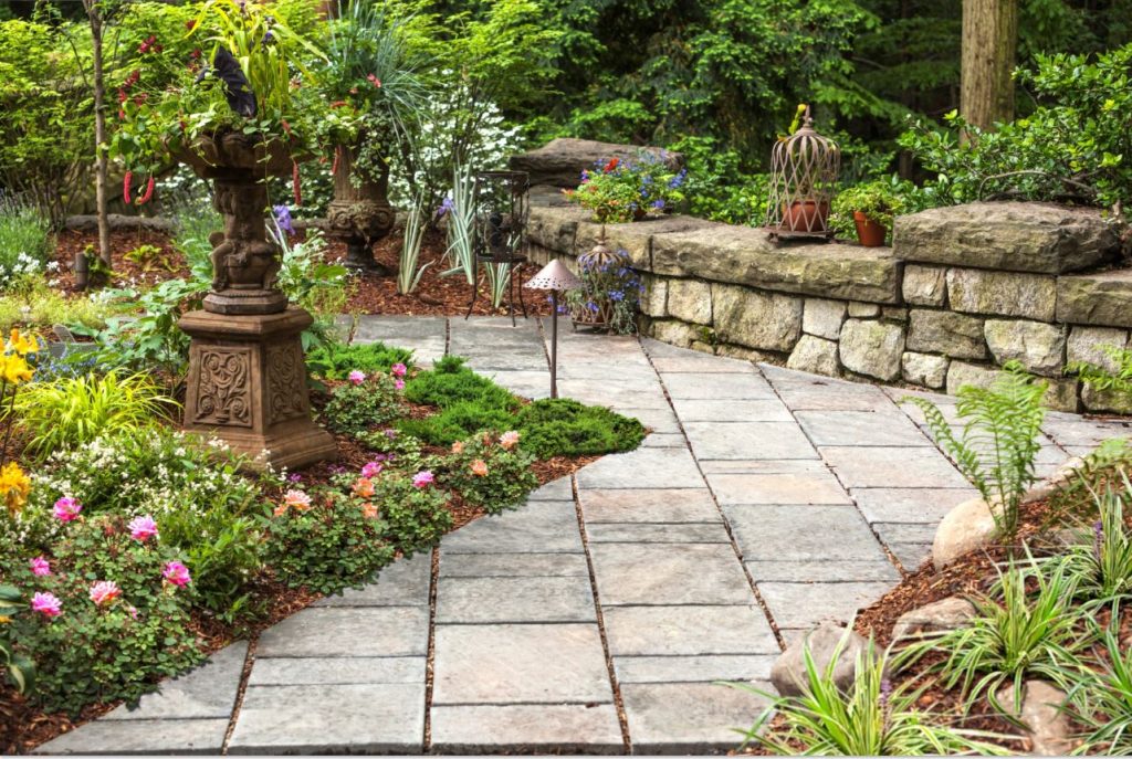 Stone garden walkway bordered by flowers, decorative planters, retaining wall, and greenery